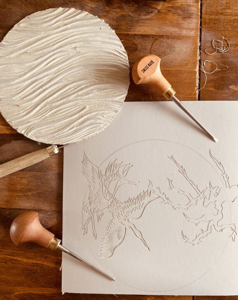 Half cut Lino of cranes flying with cut Lino circle in the background and Lino cutting tools.