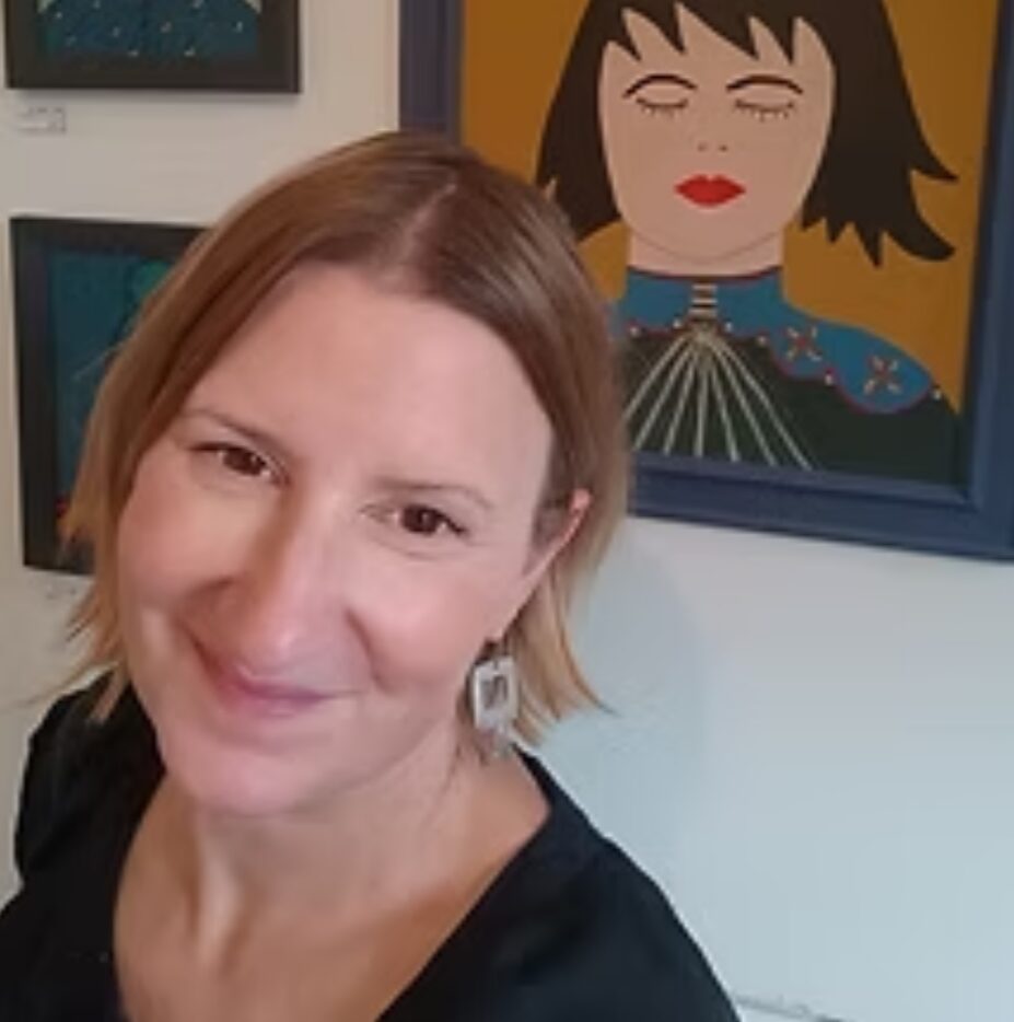 Artist Martina Munn smiling at the camera in front of one of her artworks, depicting a portrait of a lady with closed eyes, freckles,red lipstick and colourful heavily patterned top..