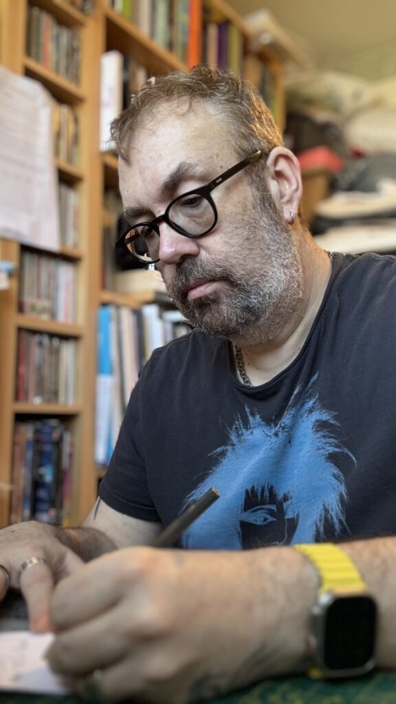 I’m seated at my desk, focused as I write with a pencil on a sheet of paper. I’m wearing glasses, a dark T‑shirt with a blue graphic, and a yellow watch. Behind me is a wall of bookshelves filled with books, sketchbooks, and materials, creating a cosy, creative studio atmosphere.